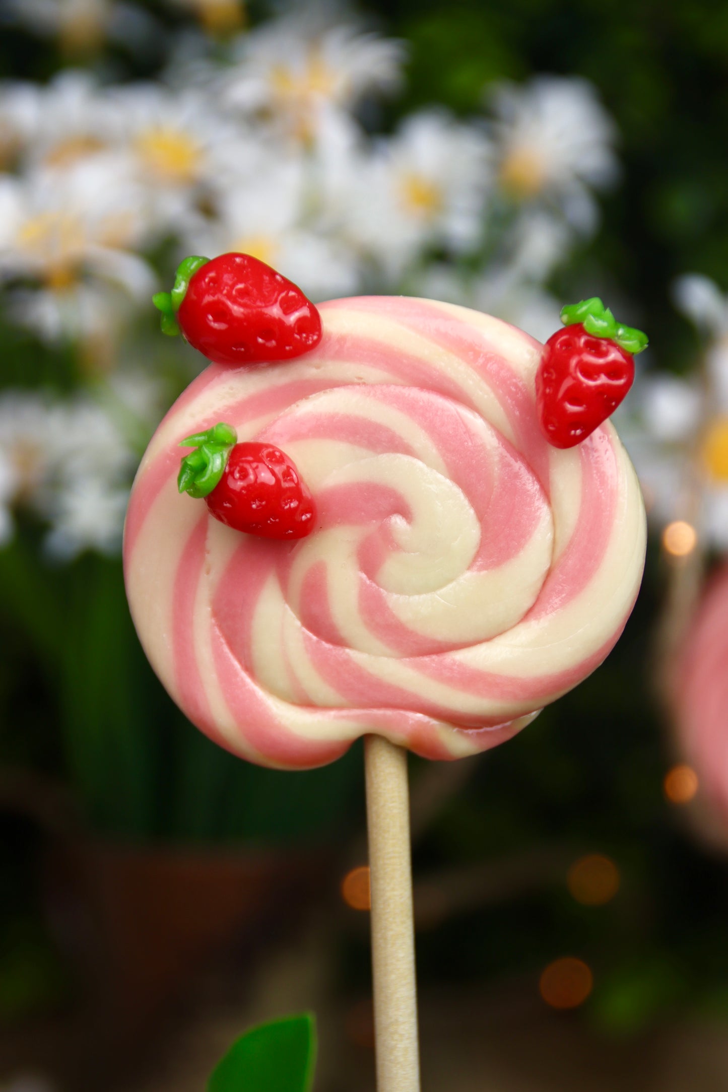 Sucette foraine artisanale en spirale, parfumée à la vanille et à la fraise, décorée de petites fraises sculptées à la main. Une gourmandise élégante et fruitée, parfaite pour le printemps.