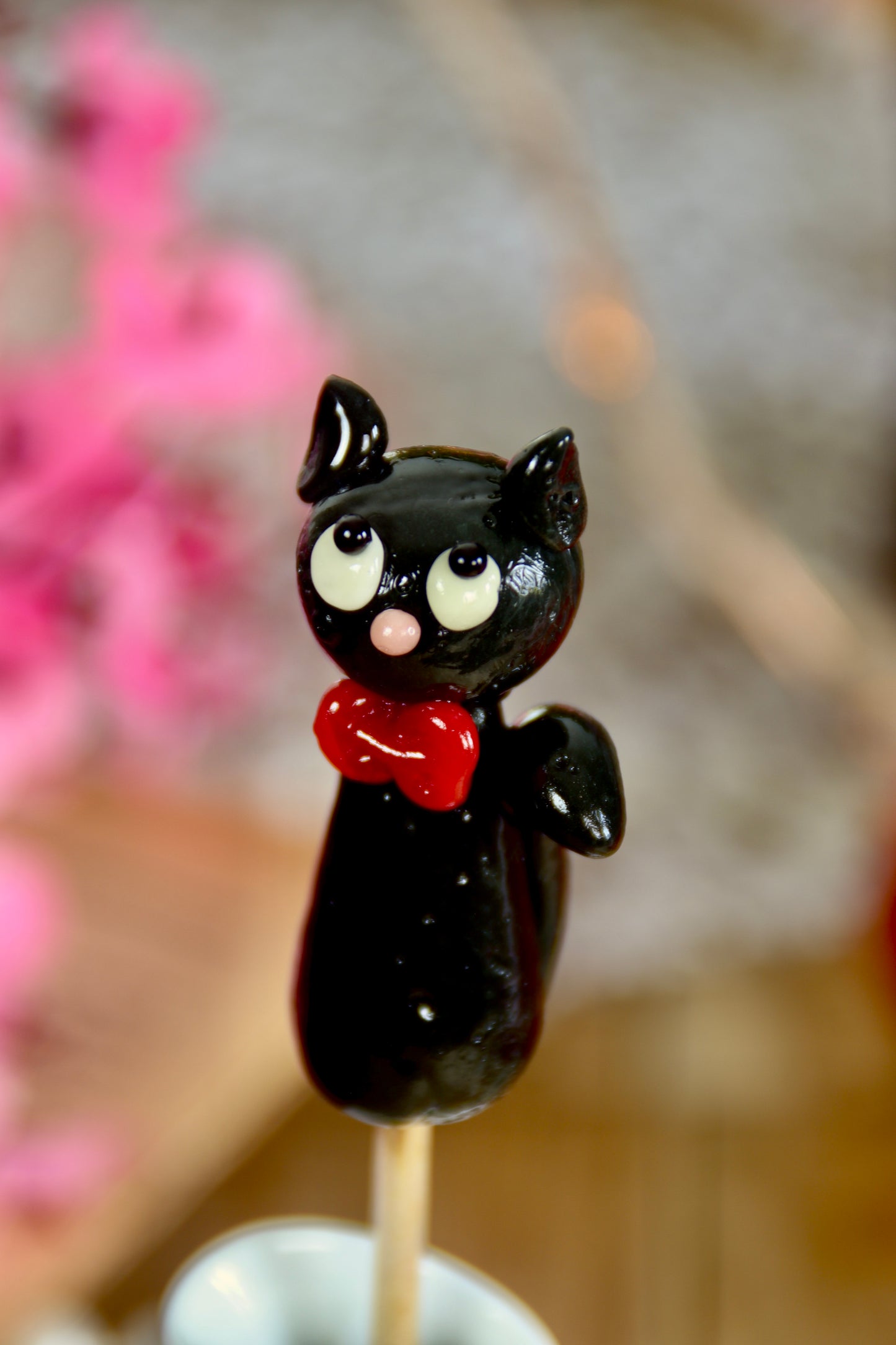 Gros plan sur une sucette noire en forme de chat avec un nœud papillon rouge, sculptée à la main et parfumée au cassis.