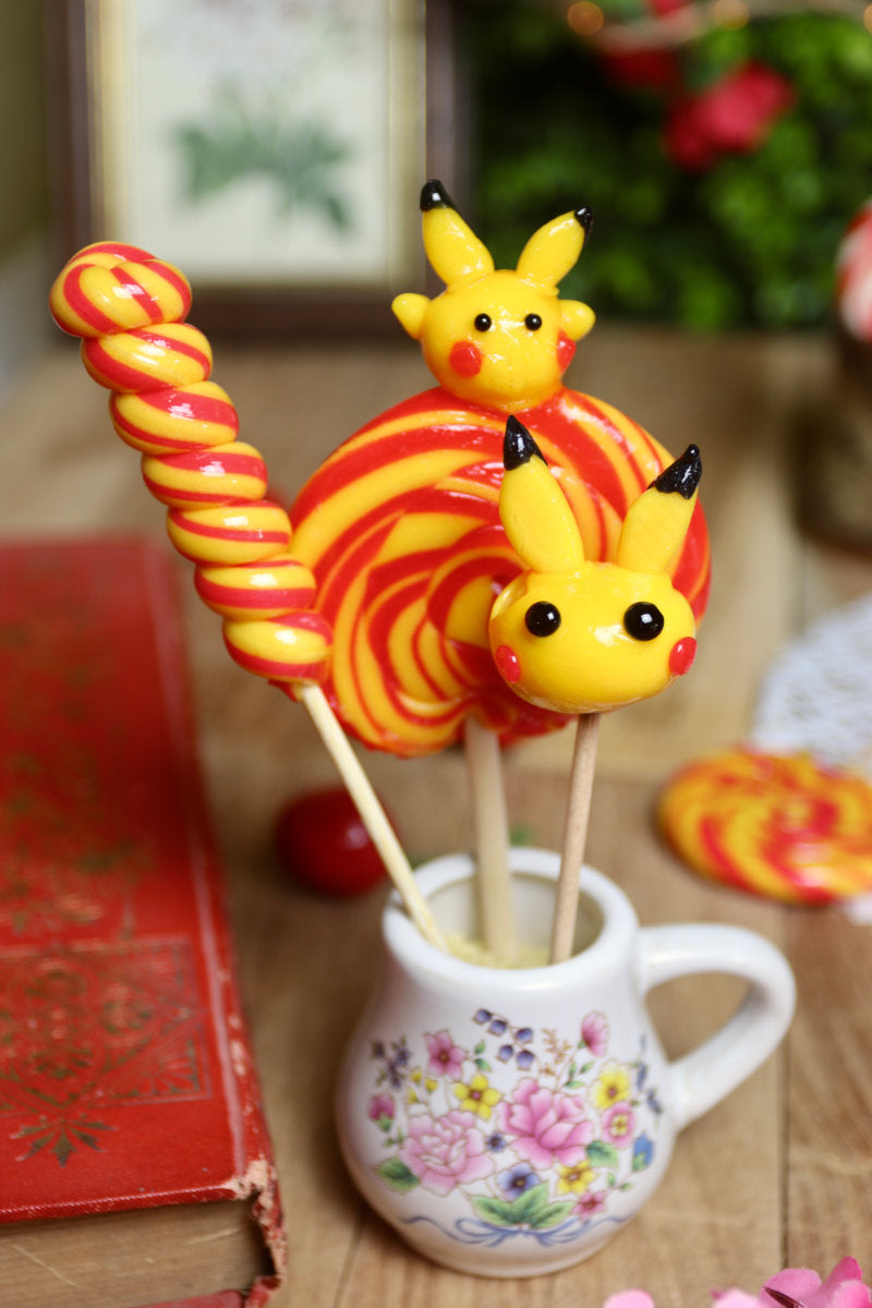 Gros plan sur les trois sucettes du lot banane-cerise : une sucette sculptée jaune inspirée de Pikachu, une torsade banane-cerise et une sucette foraine en spirale jaune et rouge.