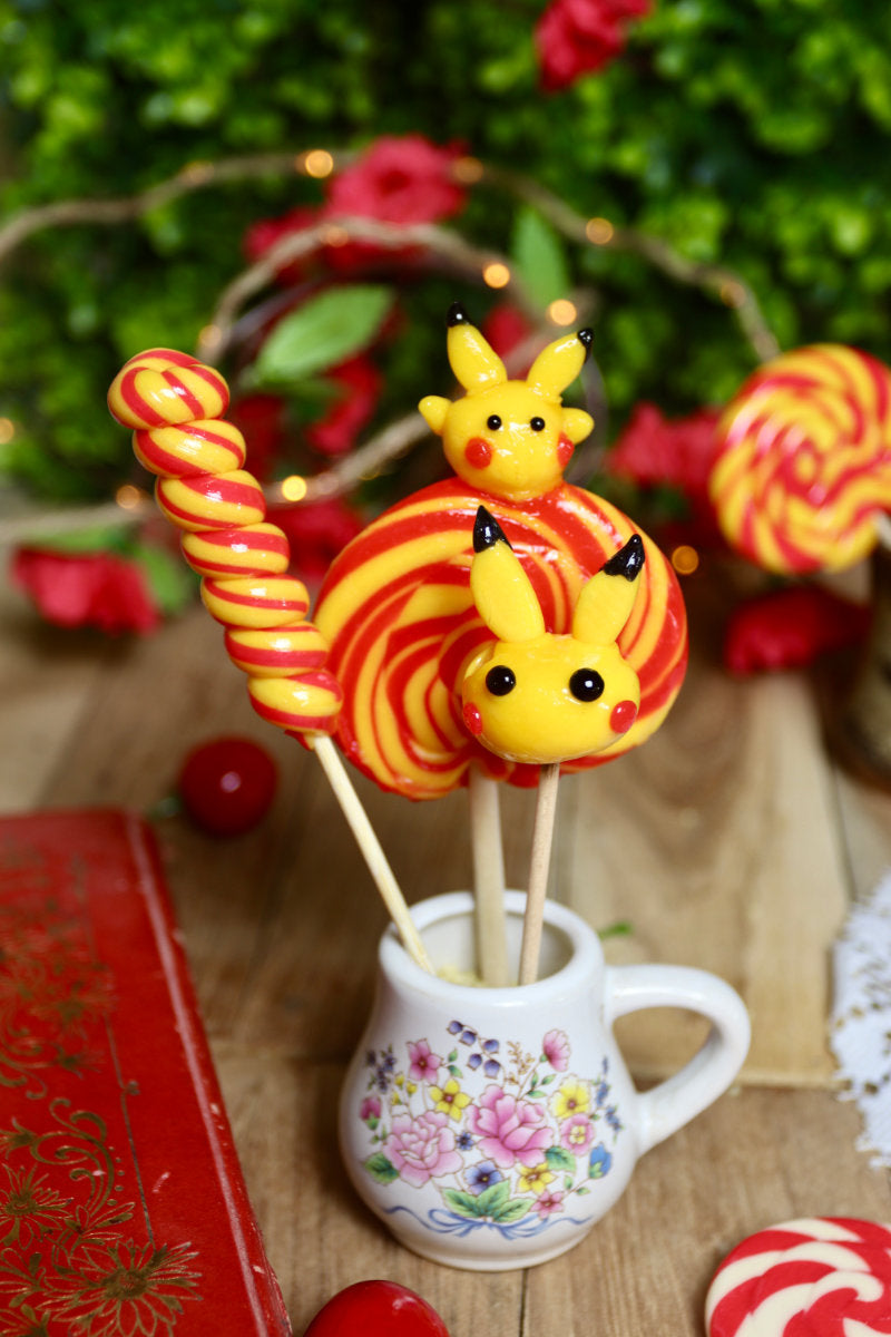 Gros plan sur les trois sucettes du lot banane-cerise : une sucette sculptée jaune inspirée de Pikachu, une torsade banane-cerise et une sucette foraine en spirale jaune et rouge.