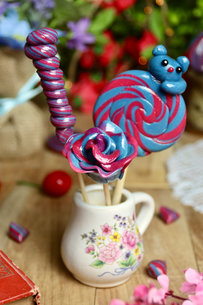 Gros plan sur les trois sucettes du lot myrtille-cerise : une spirale avec un chat sculpté, une torsade, et une rose, toutes dans les tons bleus et rouges.