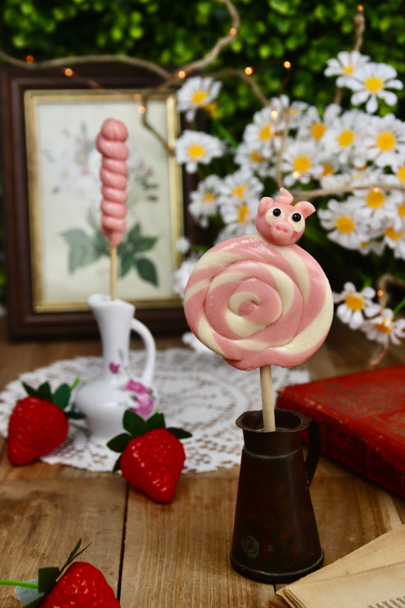 Sucette cochon vanille-fraise présentée dans un décor festif de type fête foraine, avec des accessoires gourmands et colorés