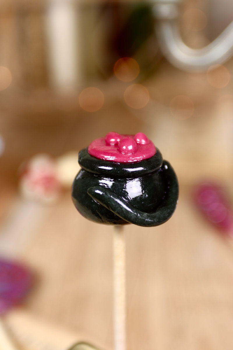 Sucette artisanale en forme de chaudron noir, sculptée à la main, avec un effet de bulles violettes à la surface – parfum cassis.