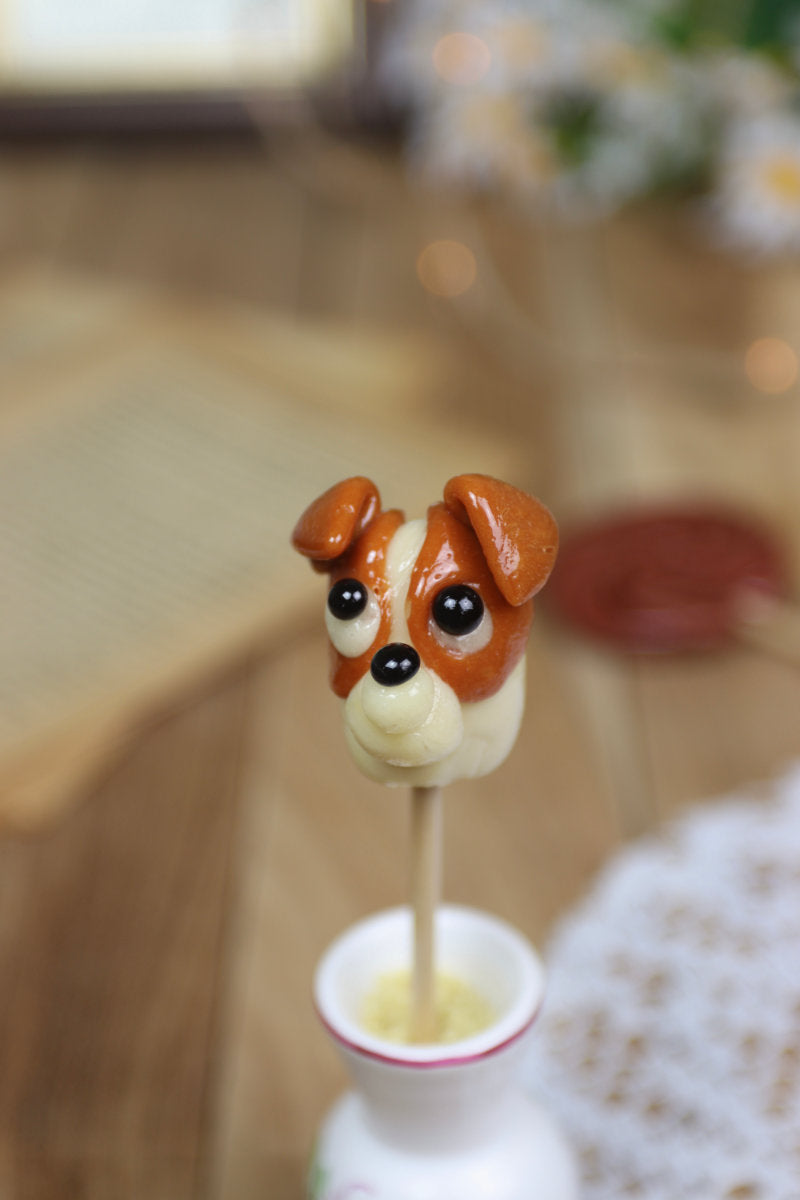 Gros plan sur une sucette artisanale en forme de Jack Russell, parfum vanille et caramel, avec détails sculptés à la main.