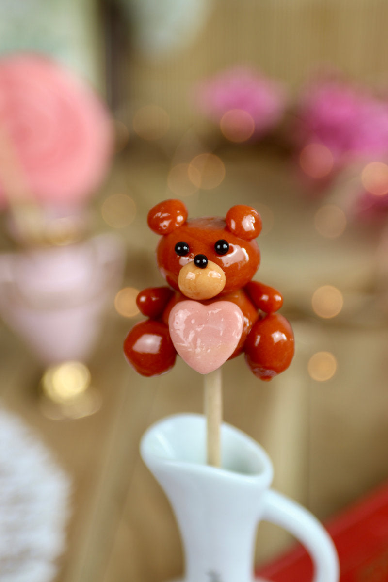 Sucette artisanale en forme de peluche au caramel avec un cœur rouge framboise sculpté sur le ventre.
