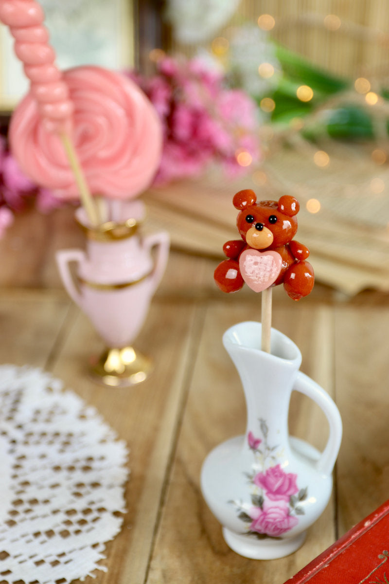 Sucette peluche posée sur un tissu doux avec lumière tamisée, ambiance cocooning et gourmande, évoquant l’enfance et les cadeaux tendres.