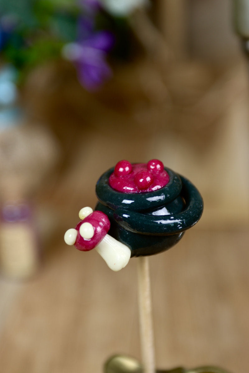 Gros plan sur une sucette artisanale en forme de chaudron au cassis, avec un petit champignon sculpté sur le rebord.