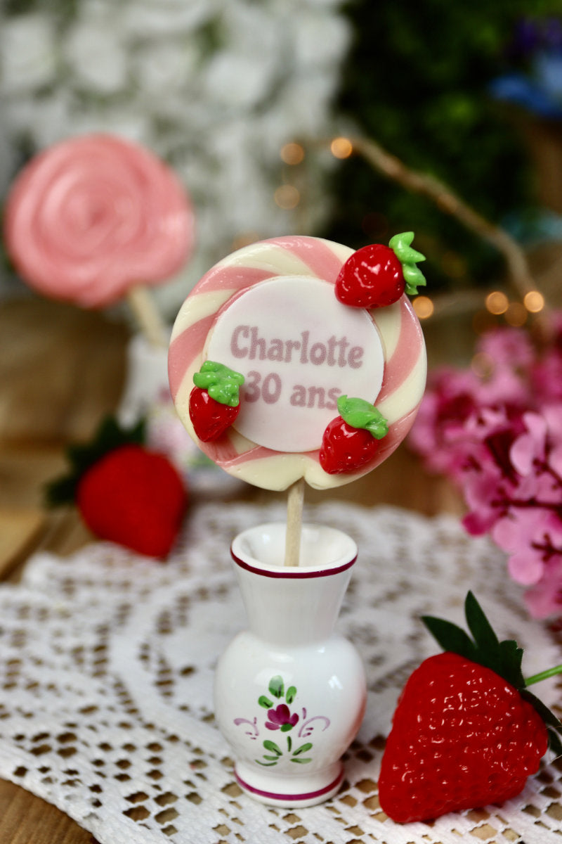 Gros plan sur une sucette spirale vanille-fraise avec petites fraises sculptées et message personnalisé au centre.