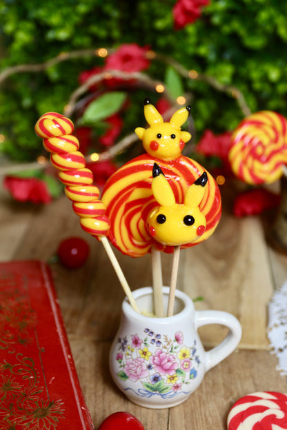 Gros plan sur les trois sucettes du lot banane-cerise : une sucette sculptée jaune inspirée de Pikachu, une torsade banane-cerise et une sucette foraine en spirale jaune et rouge.
