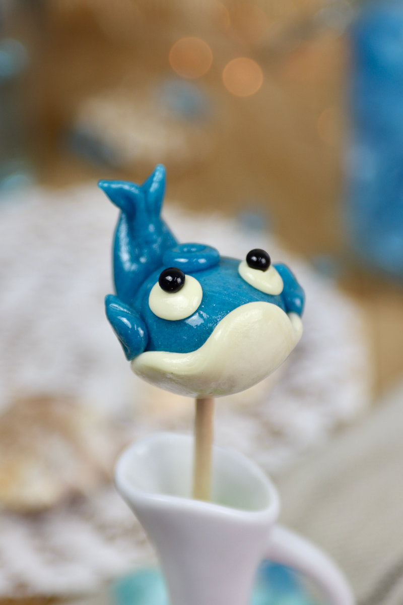 Sucette artisanale en forme de baleine, sculptée à la main, aux saveurs myrtille et citron, présentée sur un fond marin décoratif.