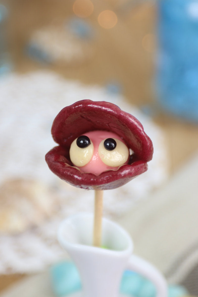Sucette artisanale en forme de coquillage, sculptée à la main, aux saveurs fraise et cassis, présentée sur un fond marin décoratif.