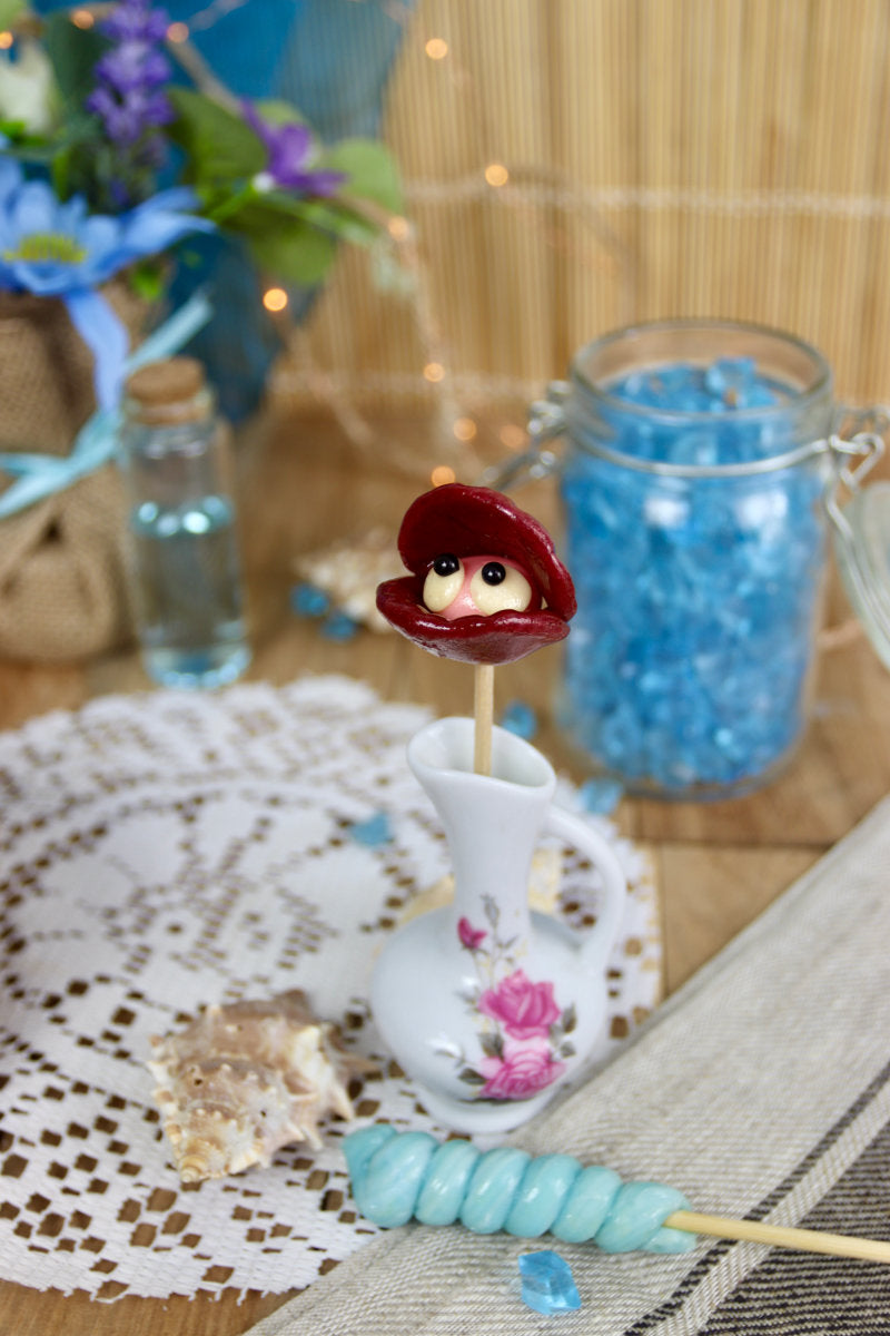Sucette coquillage fraise cassis dans un décor marin, entourée de coquillages et de sable, parfaite pour un thème océan ou une décoration gourmande.