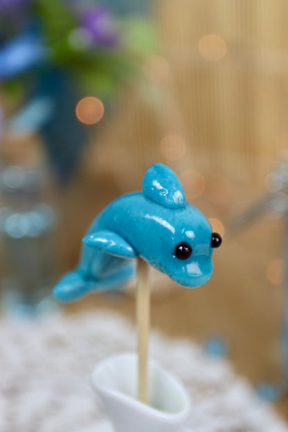 Sucette artisanale en forme de dauphin, sculptée à la main, au goût myrtille, présentée sur un fond marin décoratif.