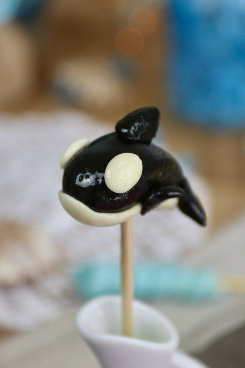 Sucette artisanale en forme d’orque, sculptée à la main, au goût vanille et cola, présentée sur un fond marin décoratif.