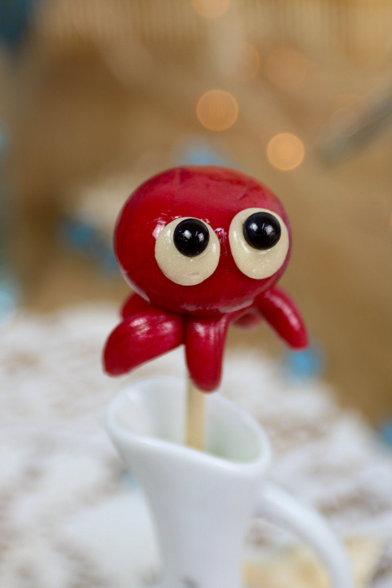 Sucette artisanale en forme de pieuvre, sculptée à la main, avec un goût cerise intense, présentée sur un fond marin décoratif.