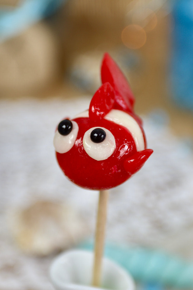 Sucette artisanale en forme de poisson, sculptée à la main, aux saveurs framboise et noix de coco, présentée sur un fond marin décoratif.