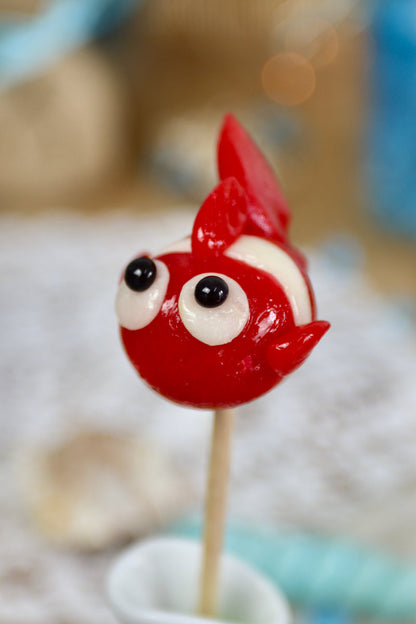 Sucette artisanale en forme de poisson, sculptée à la main, aux saveurs framboise et noix de coco, présentée sur un fond marin décoratif.