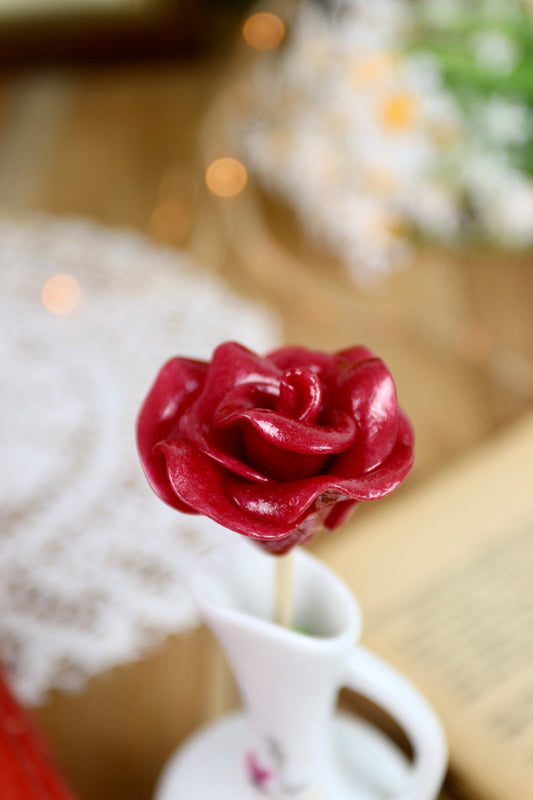Gros plan sur une sucette en forme de rose rouge, sculptée à la main, avec un parfum de cerise. Les pétales sont détaillés et réalistes.