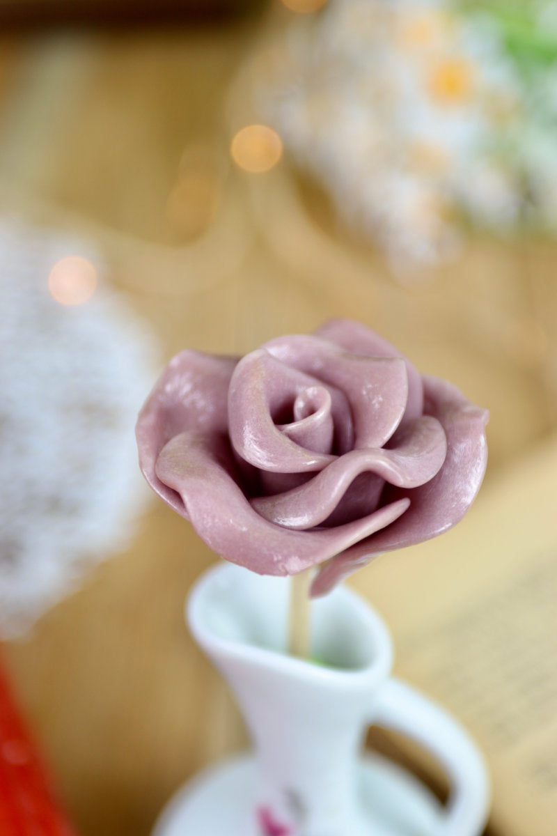 Gros plan sur une sucette artisanale en forme de rose, sculptée à la main avec un parfum de violette. Les pétales sont détaillés, rappelant la délicatesse d’une fleur fraîche.