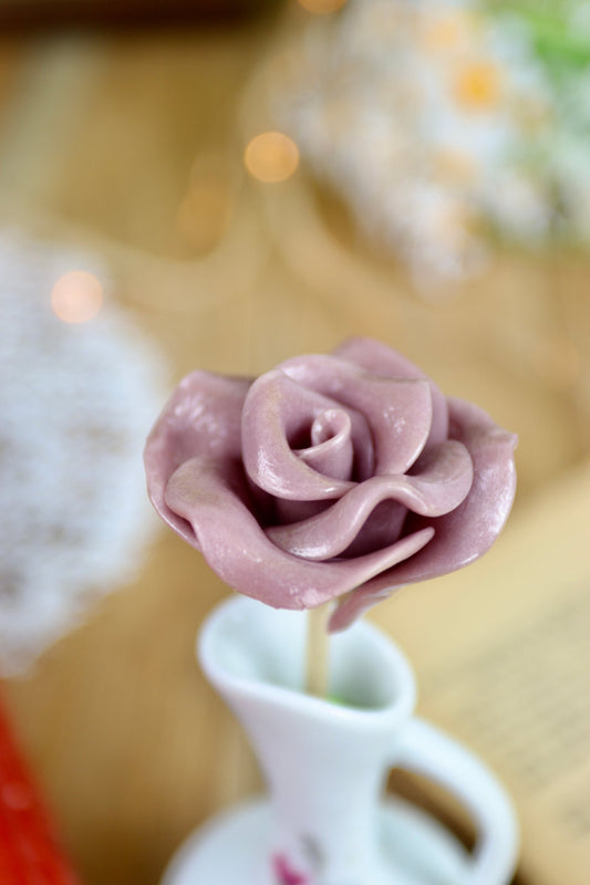 Gros plan sur une sucette artisanale en forme de rose, sculptée à la main avec un parfum de violette. Les pétales sont détaillés, rappelant la délicatesse d’une fleur fraîche.