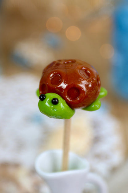 Sucette artisanale en forme de tortue, sculptée à la main, aux saveurs pomme et caramel, présentée sur un fond marin décoratif.