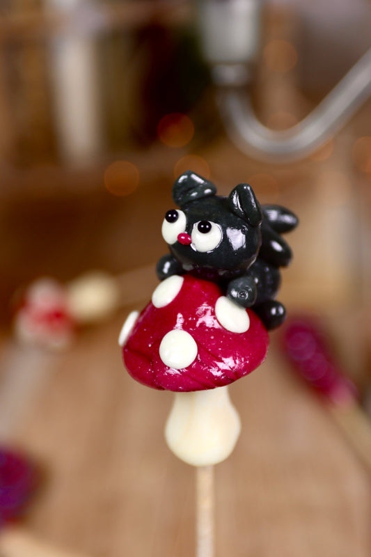 Sucette artisanale en forme de champignon vanille et cassis avec un petit chat noir sculpté à la main grimpant dessus.