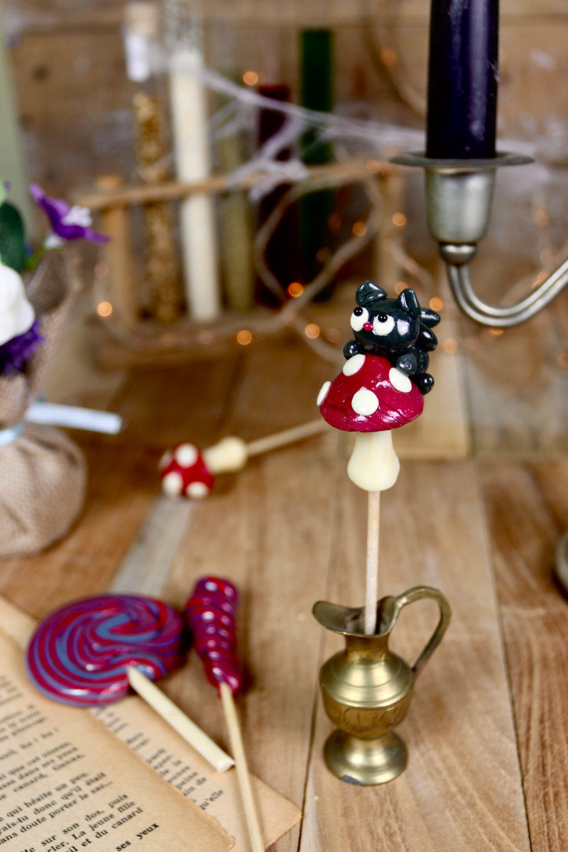 Sucette chat noir sur champignon, mise en scène dans un décor automnal féerique avec mousse, branches et lumière tamisée.