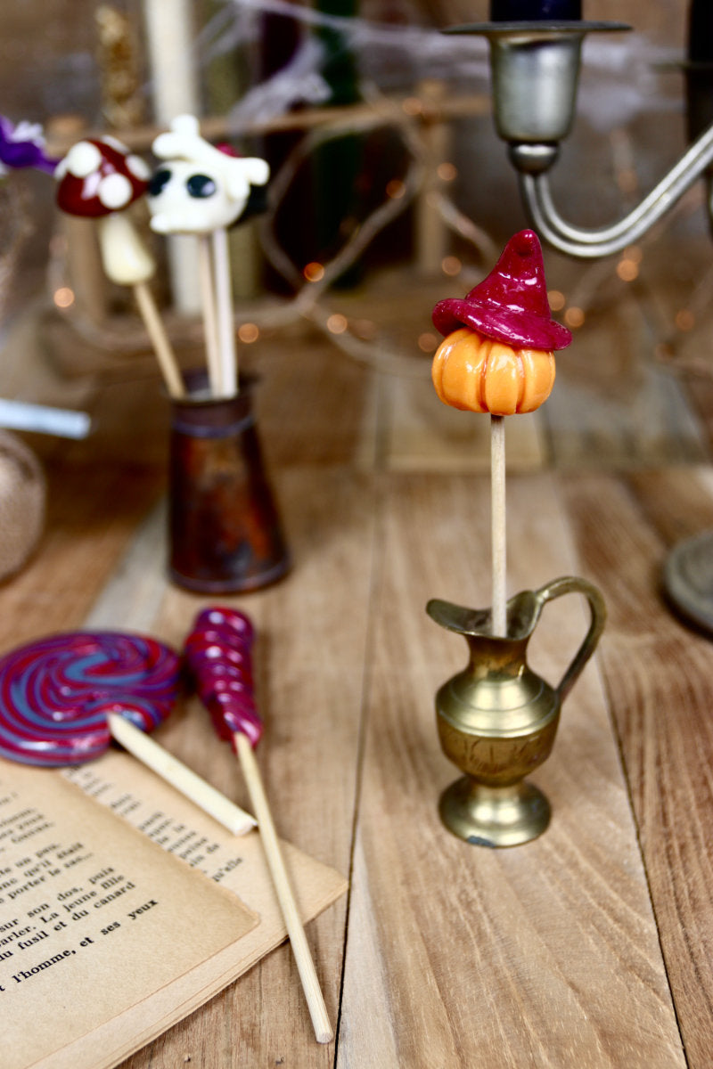 Sucette citrouille posée dans un décor automnal d’Halloween, entre feuilles, chapeaux de sorcière et chaudrons.