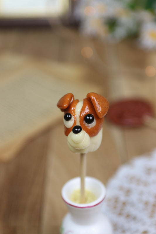 Gros plan sur une sucette artisanale en forme de Jack Russell, parfum vanille et caramel, avec détails sculptés à la main.