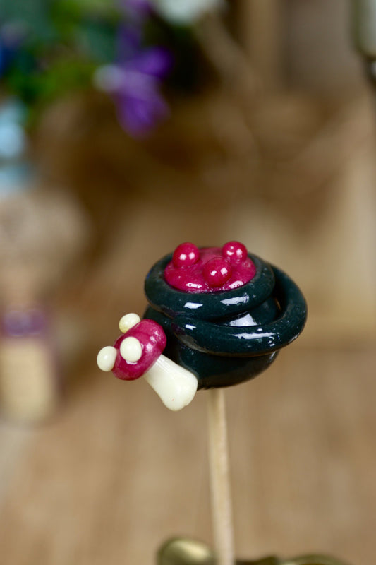 Gros plan sur une sucette artisanale en forme de chaudron au cassis, avec un petit champignon sculpté sur le rebord.