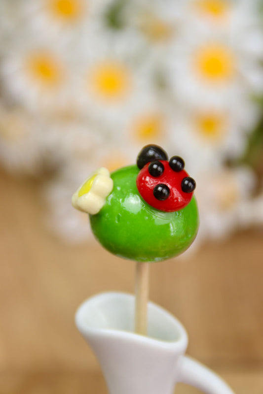 Gros plan sur une sucette verte à la pomme avec une coccinelle rouge sculptée à la main, aux détails noirs précis.