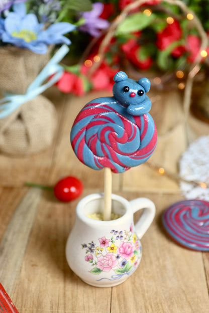 Une sucette foraine artisanale à la myrtille et à la cerise avec un chat sculptée dessus dans un décor féérique et gourmand