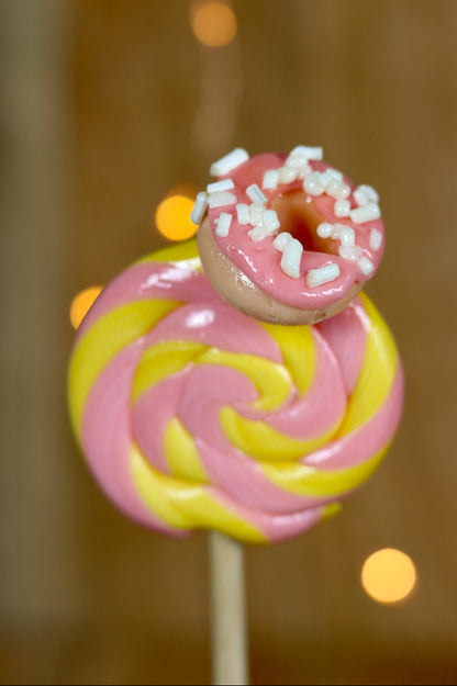 Une sucette foraine artisanale banane fraise avec un donut sculptée dessus nappé d'un glaçage fraise