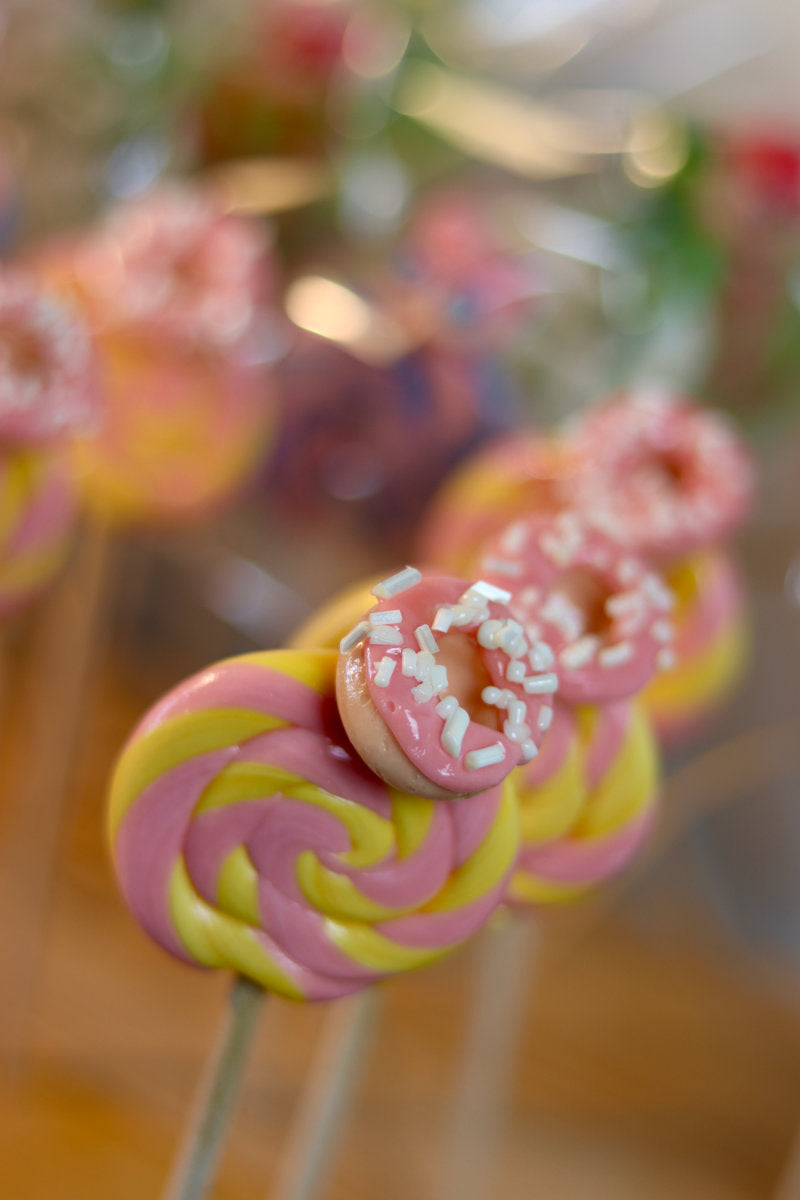 Une sucette foraine artisanale banane fraise avec un donut sculptée dessus nappé d'un glaçage fraise. dans notre atelier de farication en Bourgogne