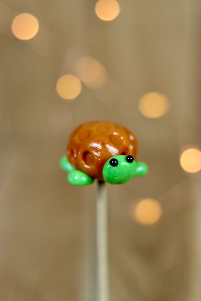 Une sucette sculptée à la main en forme de tortue. Elle est réalisée en sucre cuit aromatisé à la pomme et la carapace est au caramel. 