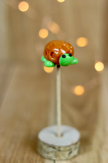 Une sucette sculptée à la main en forme de tortue. Elle est réalisée en sucre cuit aromatisé à la pomme et la carapace est au caramel. 