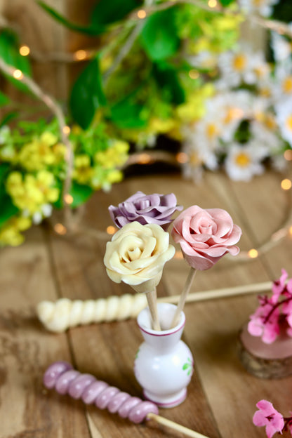 Trois Sucettes sculptées en forme de rose dans un mini vase. Une rose pêche jaune poussin, une rose framboise vieux rose et une rose à la violette. Dans un décor printanier