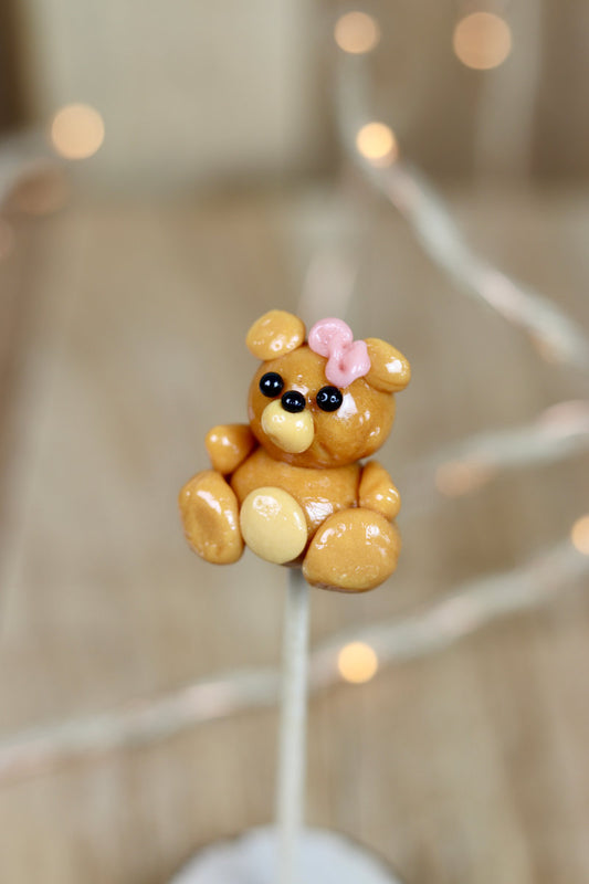 Sucette scuptée en forme de paluche au caramel beurre salé. Elle a un petit noeud papillon sur la tête rose à la fraise