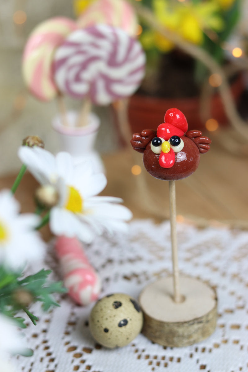 Sucette en sucre cuit sculptée en forme de poule au cacao et à la framboise. Dans un décor printanier