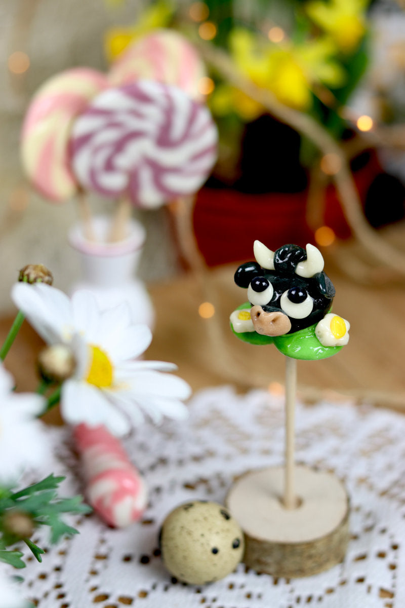 Sucette sculptée en forme de taureau dans une prairie à la pomme entouré de pâquerettes. En sucre cuit.  Dans un décor printanier