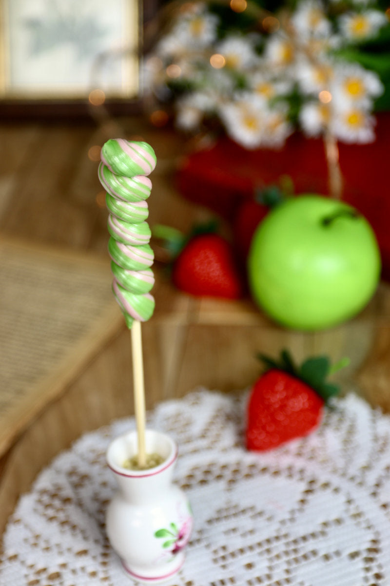 Sucette torsade pomme-fraise posée dans une ambiance féerique et naturelle, évoquant une confiserie d’antan.