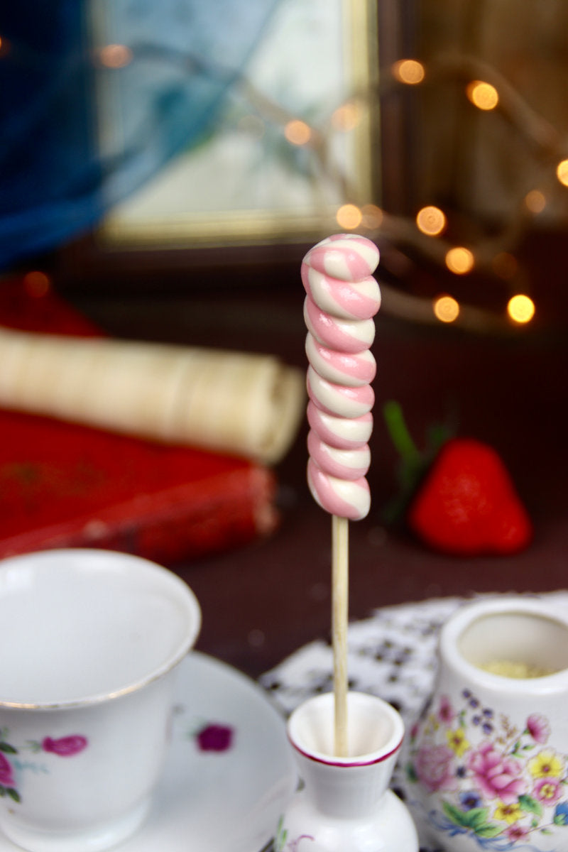 Sucette torsade Vanille-Fraise mise en scène dans un décor féerique et pastel, entourée de fleurs et de rubans, ambiance vintage et gourmande