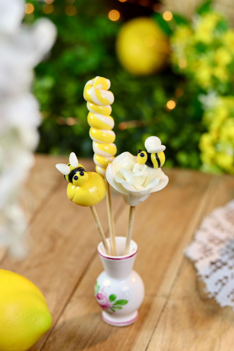 Une lot de sucette au citron. Il y a une sucette sculptée en forme d'abeille, une rose blanche avec une abeille en sucre cuit sculptée dessus et une sucette torsade citron. Dans un décor magique au citron