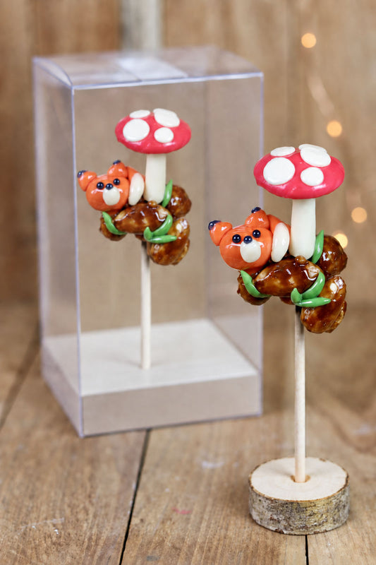 Une sucette sculptée en forme de renard sous un champignon, se reposant sur des noisettes et amandes grillés. A la pêche et à la cerise