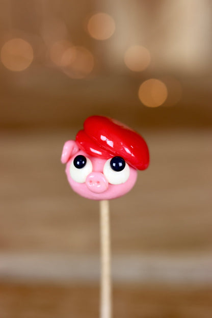 Une sucette sculptée en forme de cochon portant un béret rouge à la fraise. Photo prise en gros plan