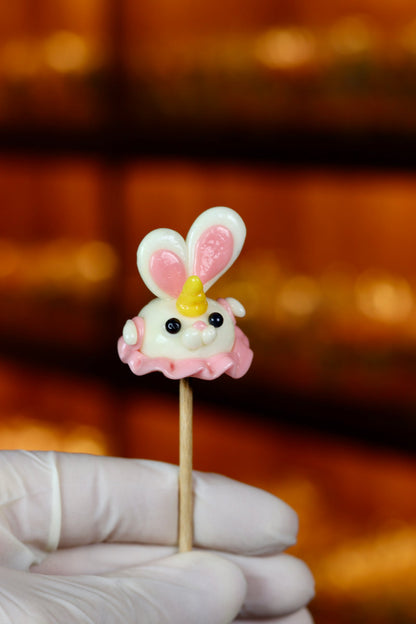 Une sucette sculptée en sucre cuit. Elle est en forme de lapin à la banane, qui porte une petite robe rose à la fraise et une corne sur le front. Photo prise dans la boutique dans un univers magique