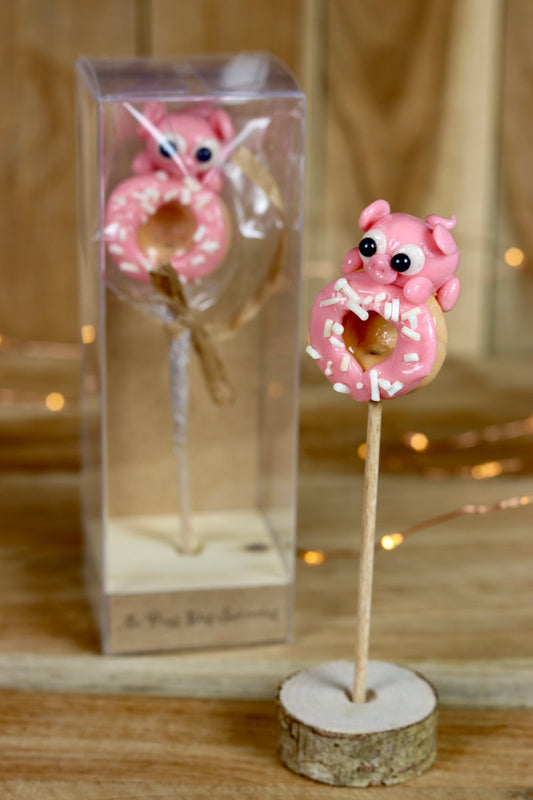 Une sucette sculptée en forme de cochon à la fraise qui grimpe sur un donut à la vanille et à la fraise. Vendu dans une boite de collection.  